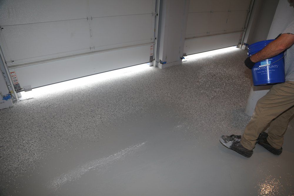 A person applying a gray epoxy coating to a garage floor from a blue bucket. The garage door is partially open.