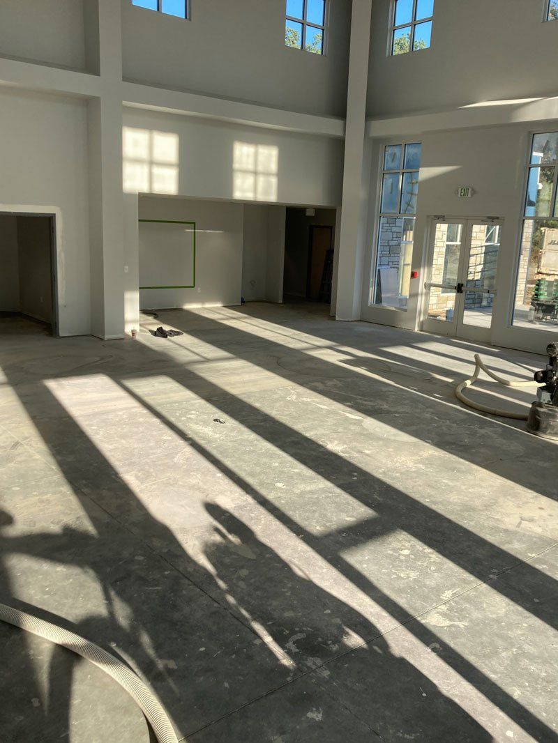 Interior view of a spacious, light-filled room under construction. Concrete floor, tall windows.