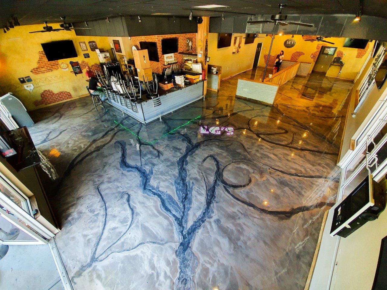 Interior view of a bar with a decorative metallic epoxy floor resembling a tree. The walls are yellow and adorned with TVs.