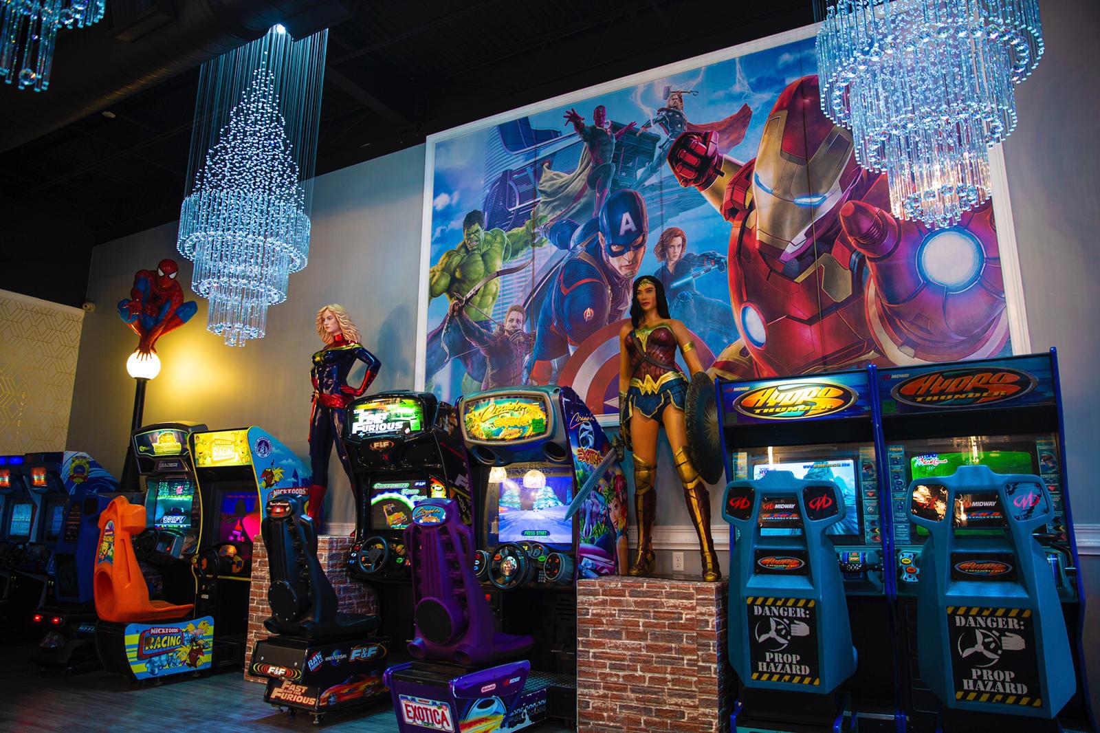 Arcade room with superhero murals and rows of colorful game machines under blue hanging lights