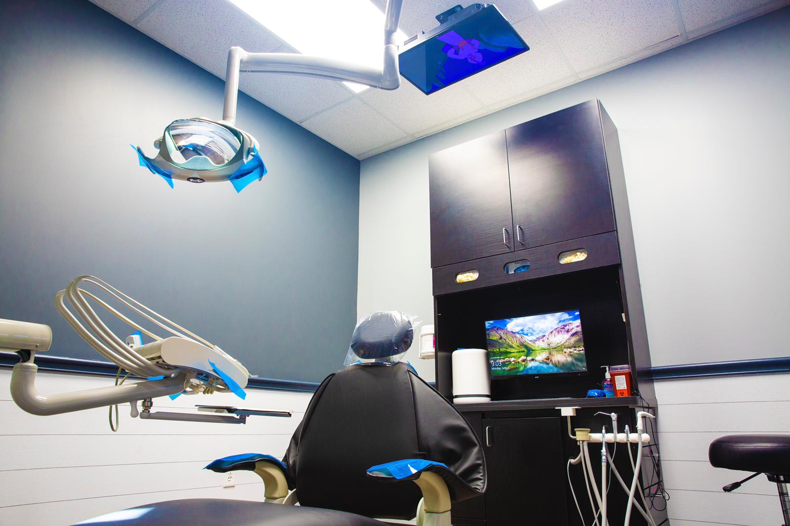 Dental clinic room with a patient chair, overhead lights, and a monitor showing a landscape scene.