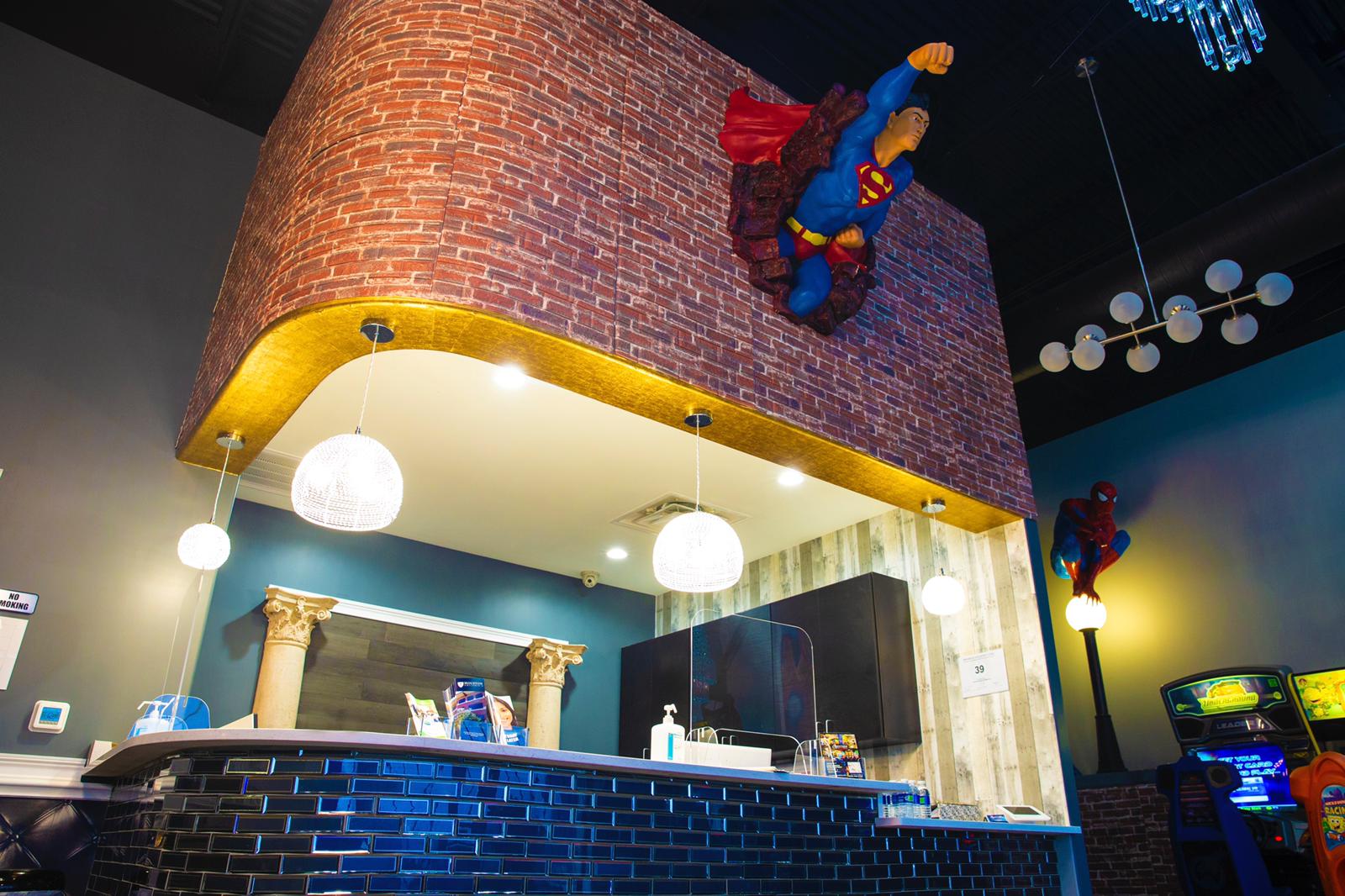 Colorful indoor scene with a curved brick wall, hanging globe lights, and superhero statues above a bar counter