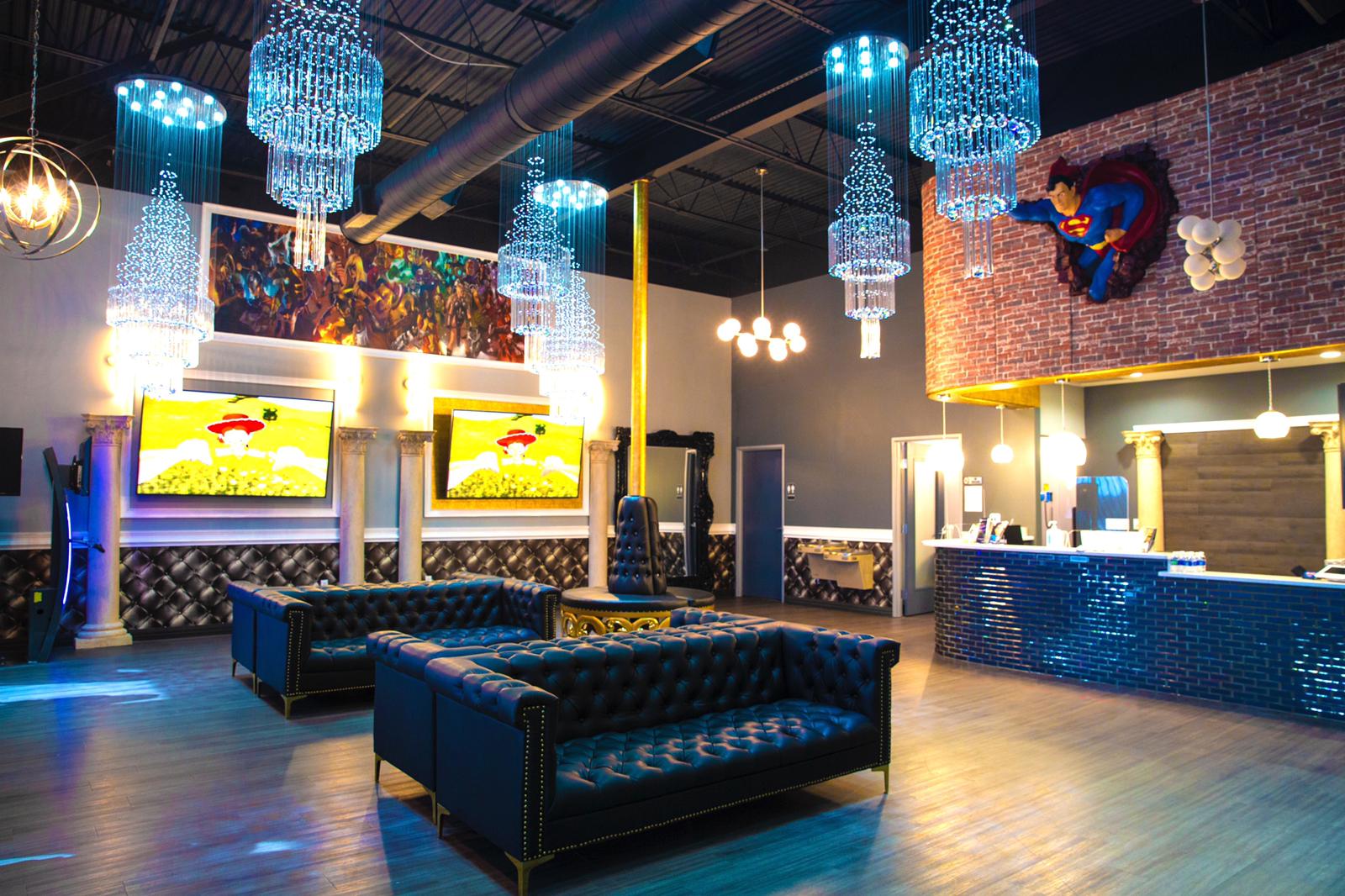 Modern lounge with blue tufted sofas, glowing chandeliers, wall TVs, and a lit bar counter