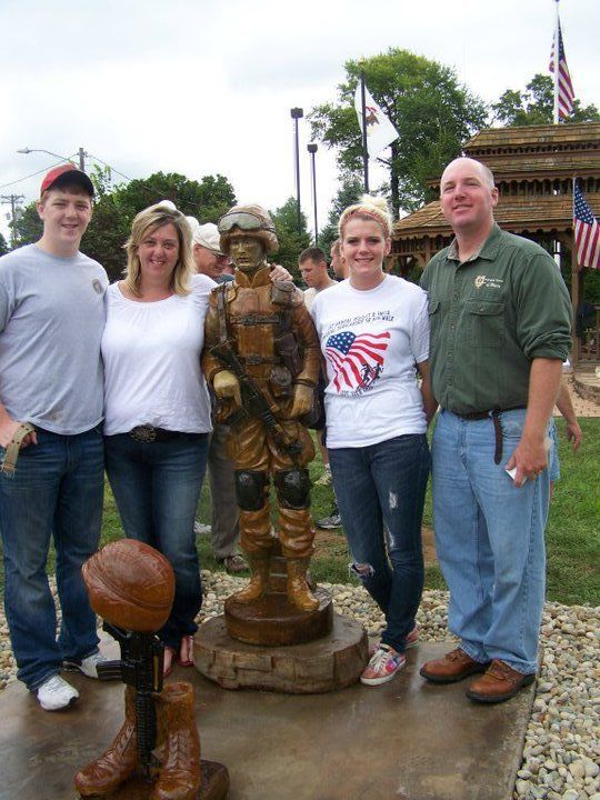 Family posing with a bronze soldier statue in an outdoor memorial setting, with American flags visible nearby.