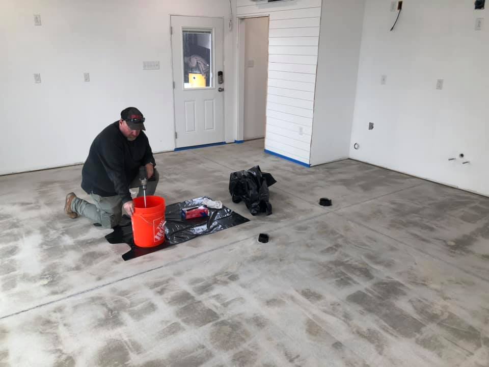 Worker kneeling on a dusty floor in an empty room, near a red bucket and tools.