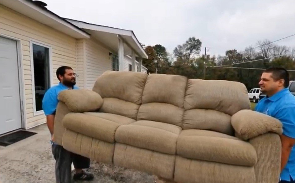 Two men are carrying a couch in front of a house.