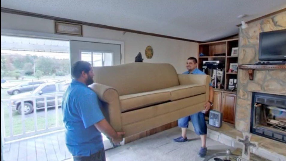 Two men are carrying a couch in a living room.