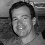 Smiling man in a dark shirt posing indoors in front of a textured wall and plants