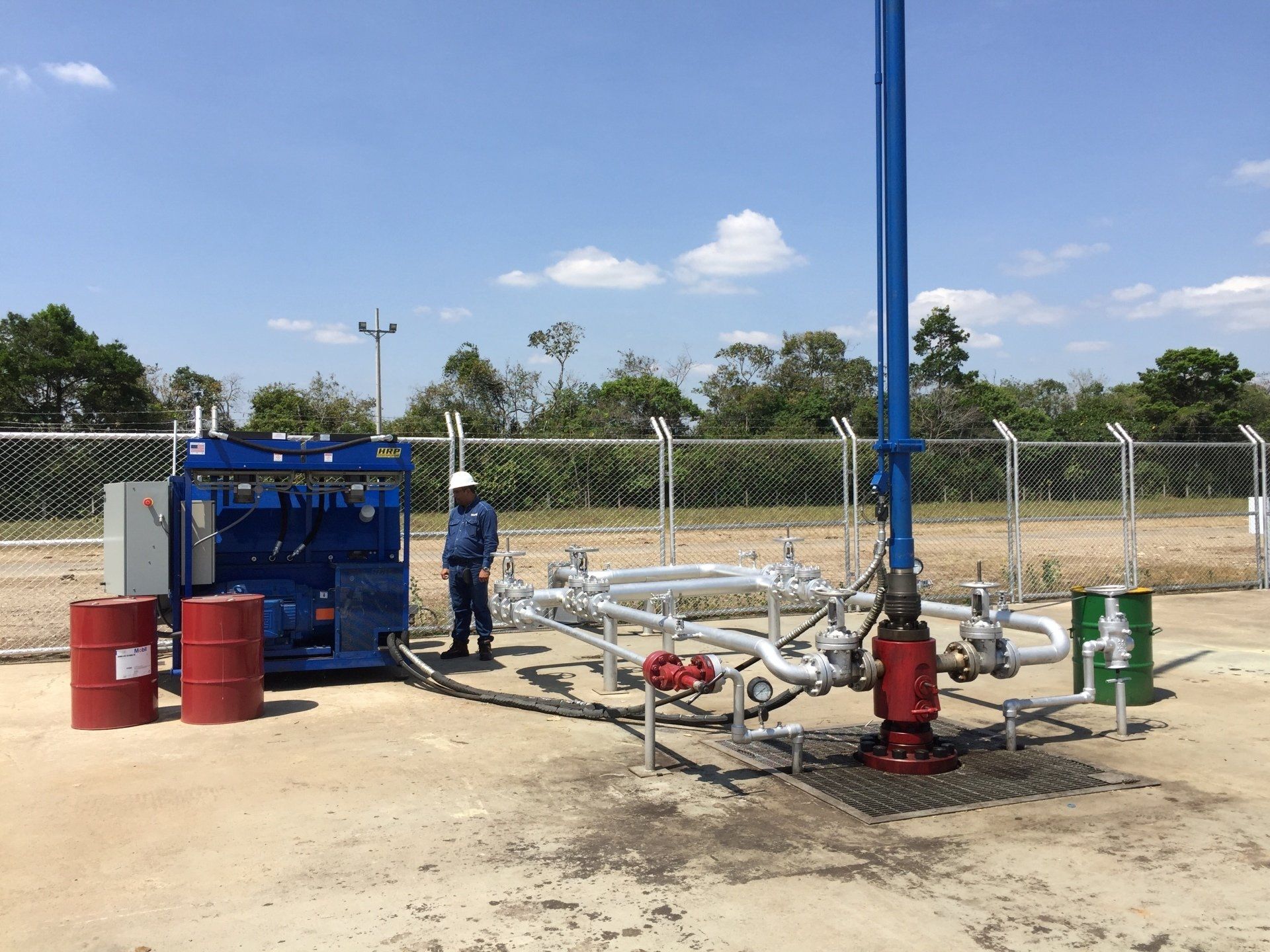 Oil/gas well site with blue equipment, pipes, red drums, and a worker outdoors under a clear sky