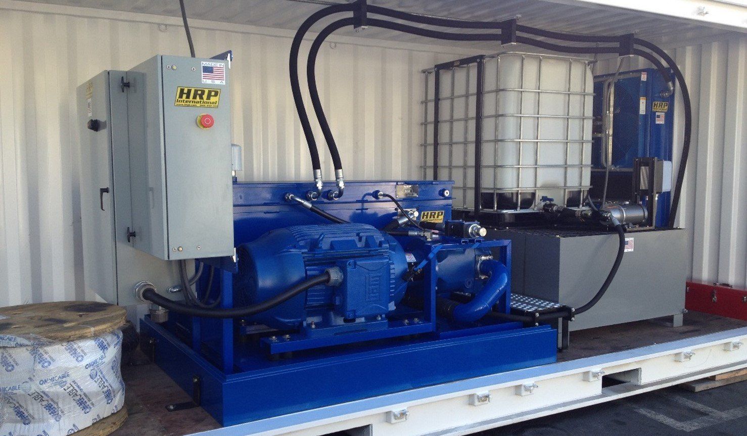 Blue industrial hydraulic machine on a metal platform with hoses, control box, and large white tank in a workshop