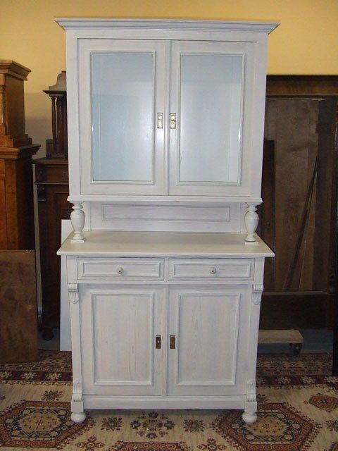 Weißer Holzschrank mit Glastüren, der auf einem gemusterten Teppich im Innenbereich steht.