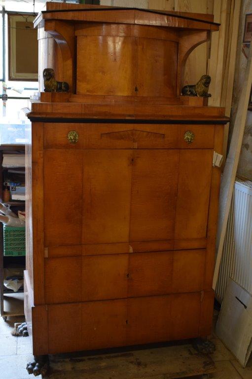 Ein antiker Holzschrank mit geschwungener Oberseite steht in einem vollgestellten Raum.