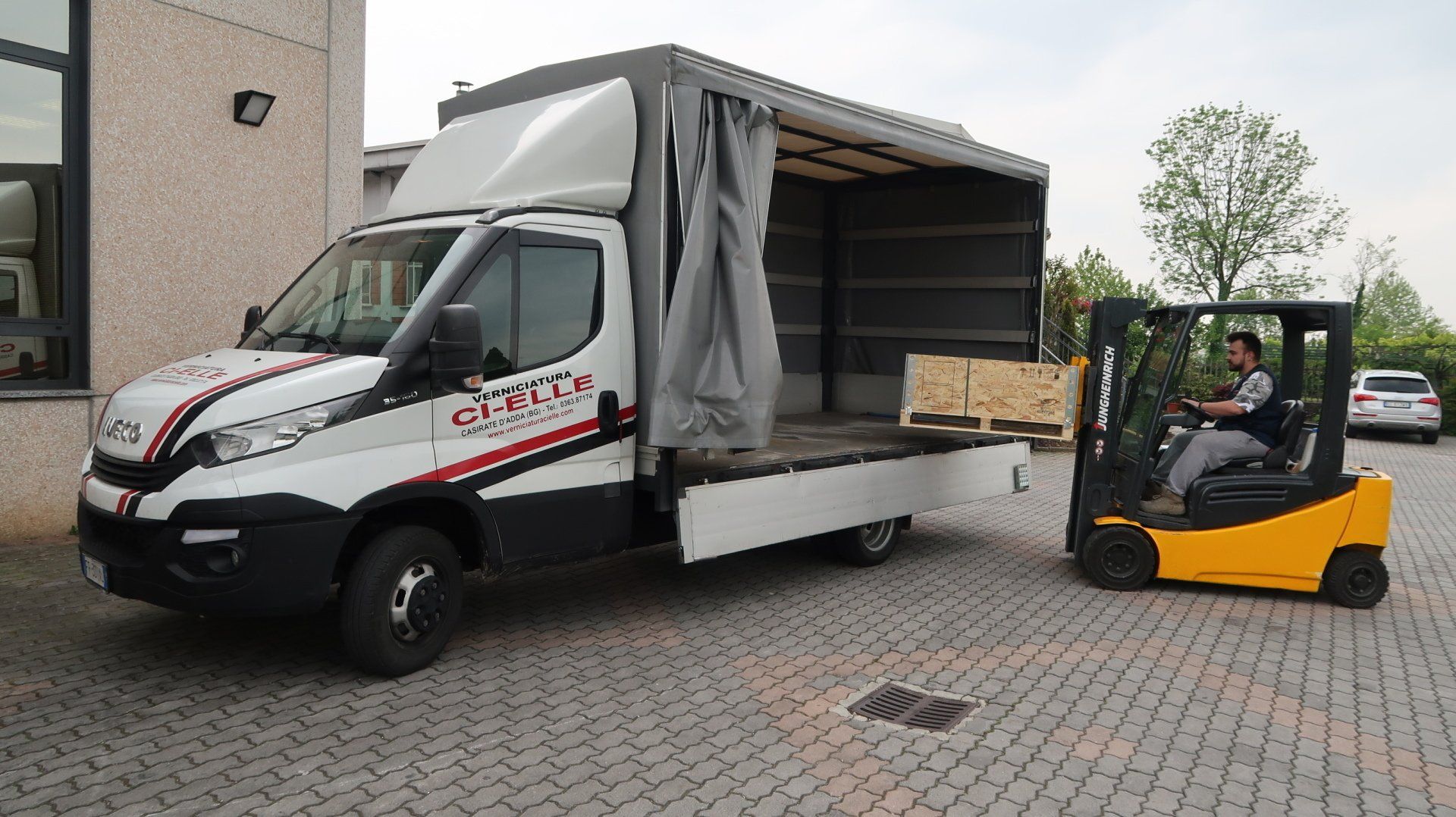 Un camioncino bianco con cassone aperto lateralmente viene caricato da un carrello elevatore giallo in un parcheggio.