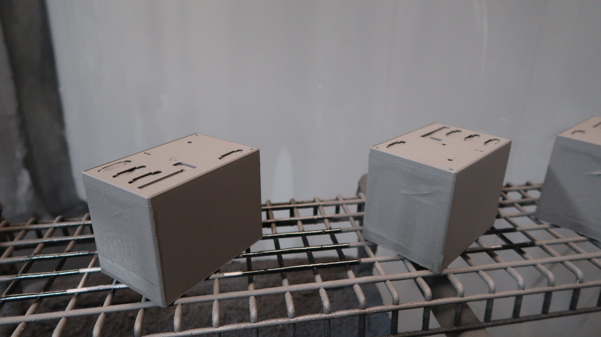 Due piccoli blocchi grigi a forma di cubo, con fori sulla parte superiore, su una griglia metallica, in un ambiente simile a un'officina.