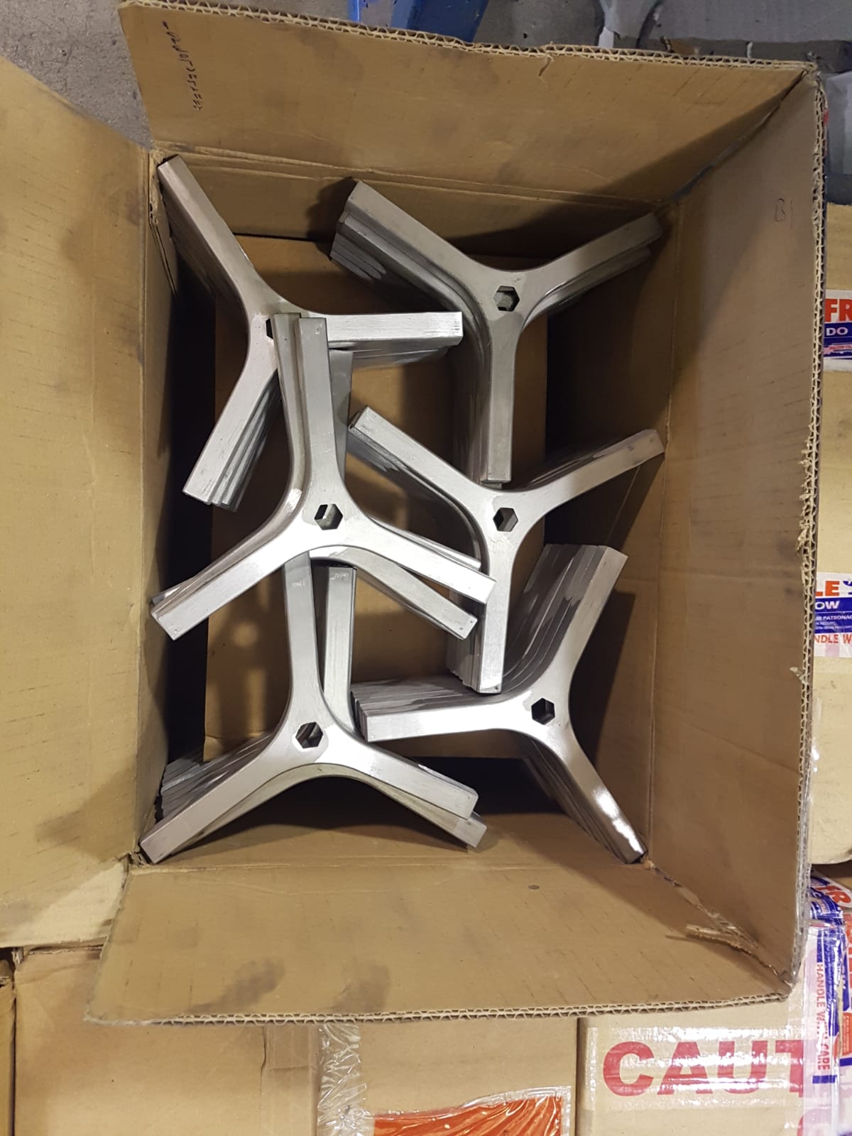 Una scatola di cartone piena di staffe metalliche argentate a tre gambe impilate sul pavimento di un magazzino.