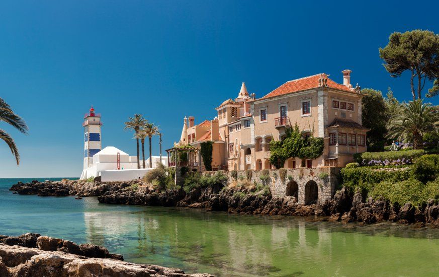 There is a lighthouse and a large house on the shore of the ocean.