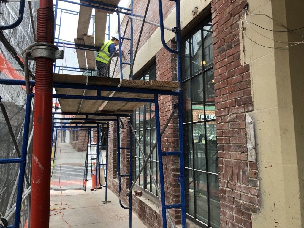 Construction worker on scaffolding, repairing brick building exterior. Red brick, large windows, blue scaffolding, safety vest.