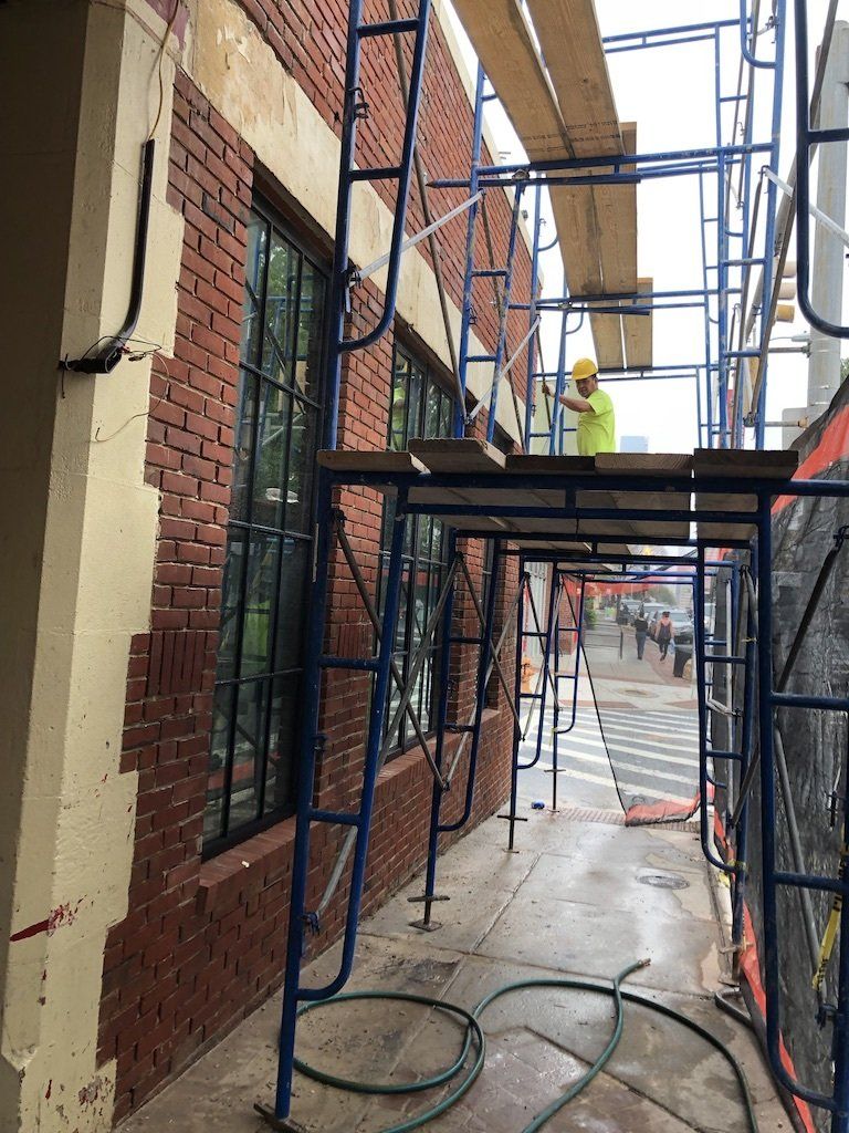 Scaffolding along a brick building. A worker in a yellow vest is on the platform. Sidewalk and street view.