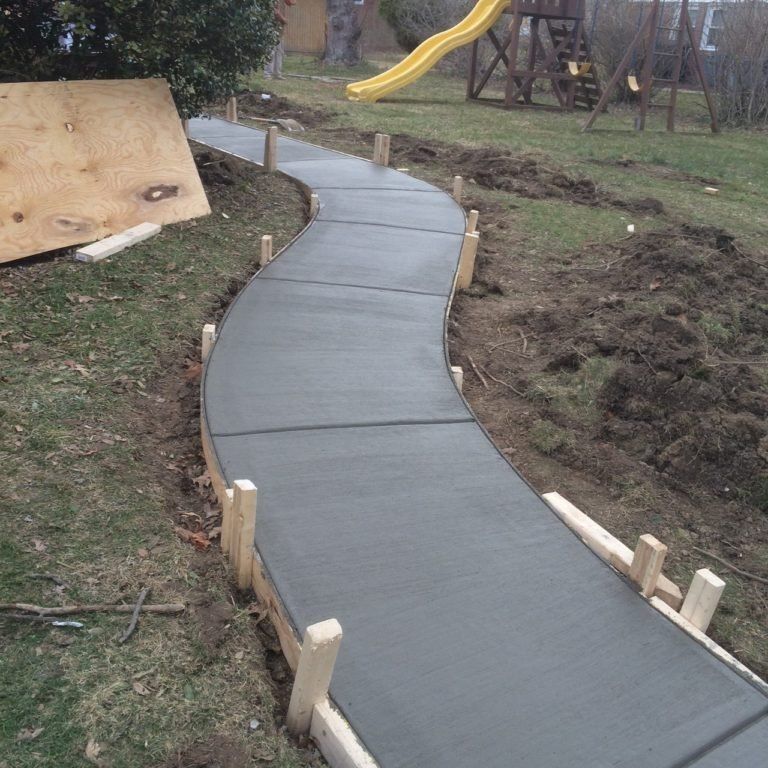 Newly poured concrete pathway with wooden forms in a grassy backyard, curving toward a playground.