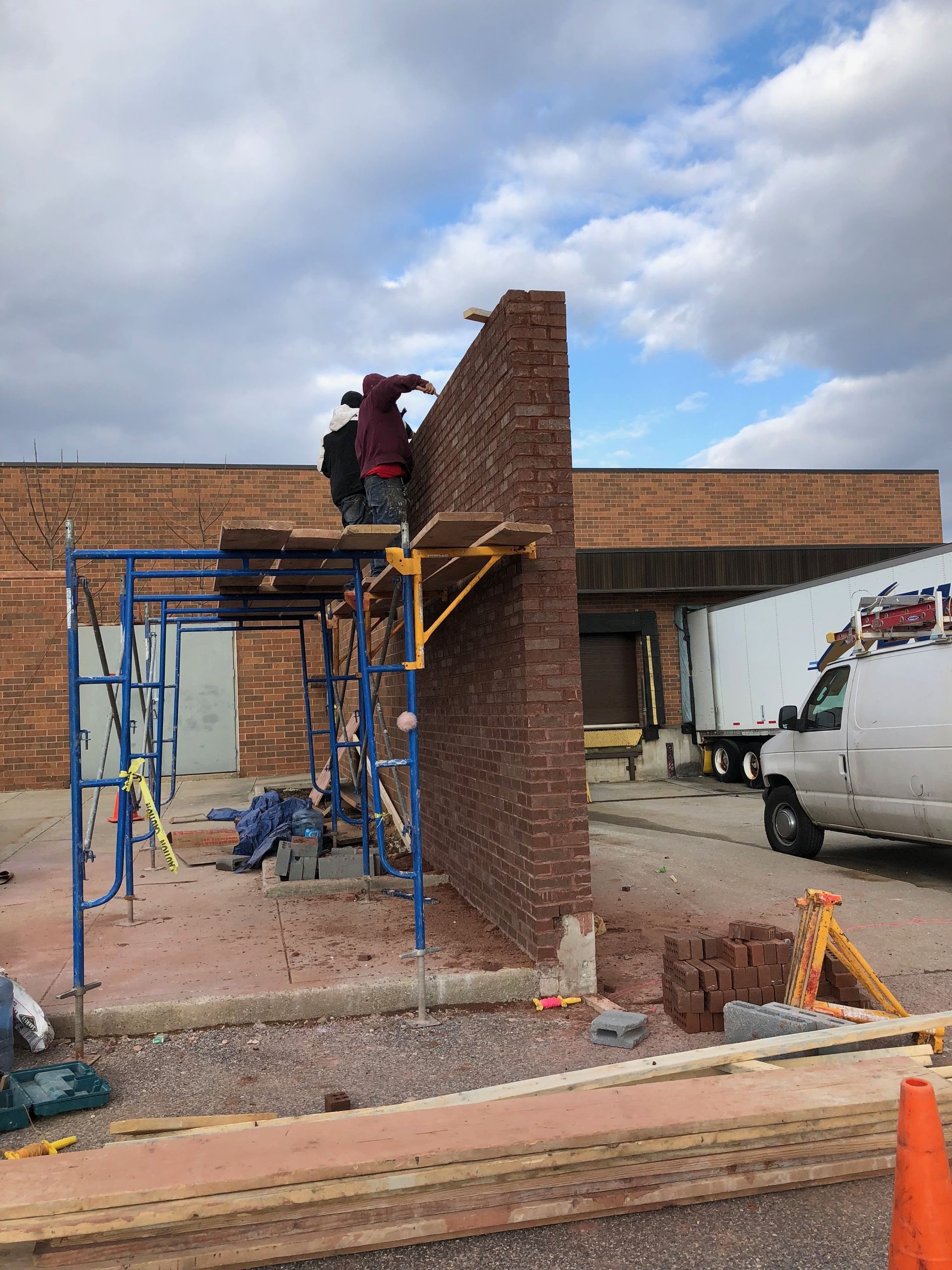 Construction worker on scaffolding laying brick wall; exterior setting. Blue sky, white truck, red brick.