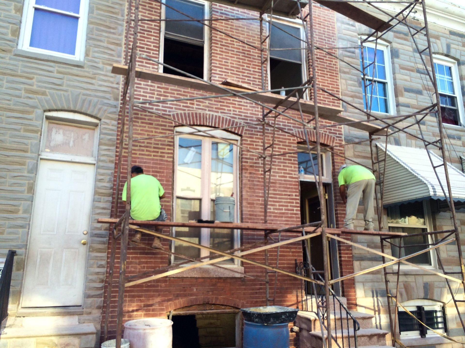 Two workers on scaffolding, repairing brick facade of a row house. One is sitting, one is standing.