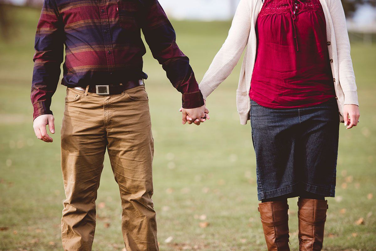 A couple stands in a grassy field holding hands, wearing casual autumn attire.
