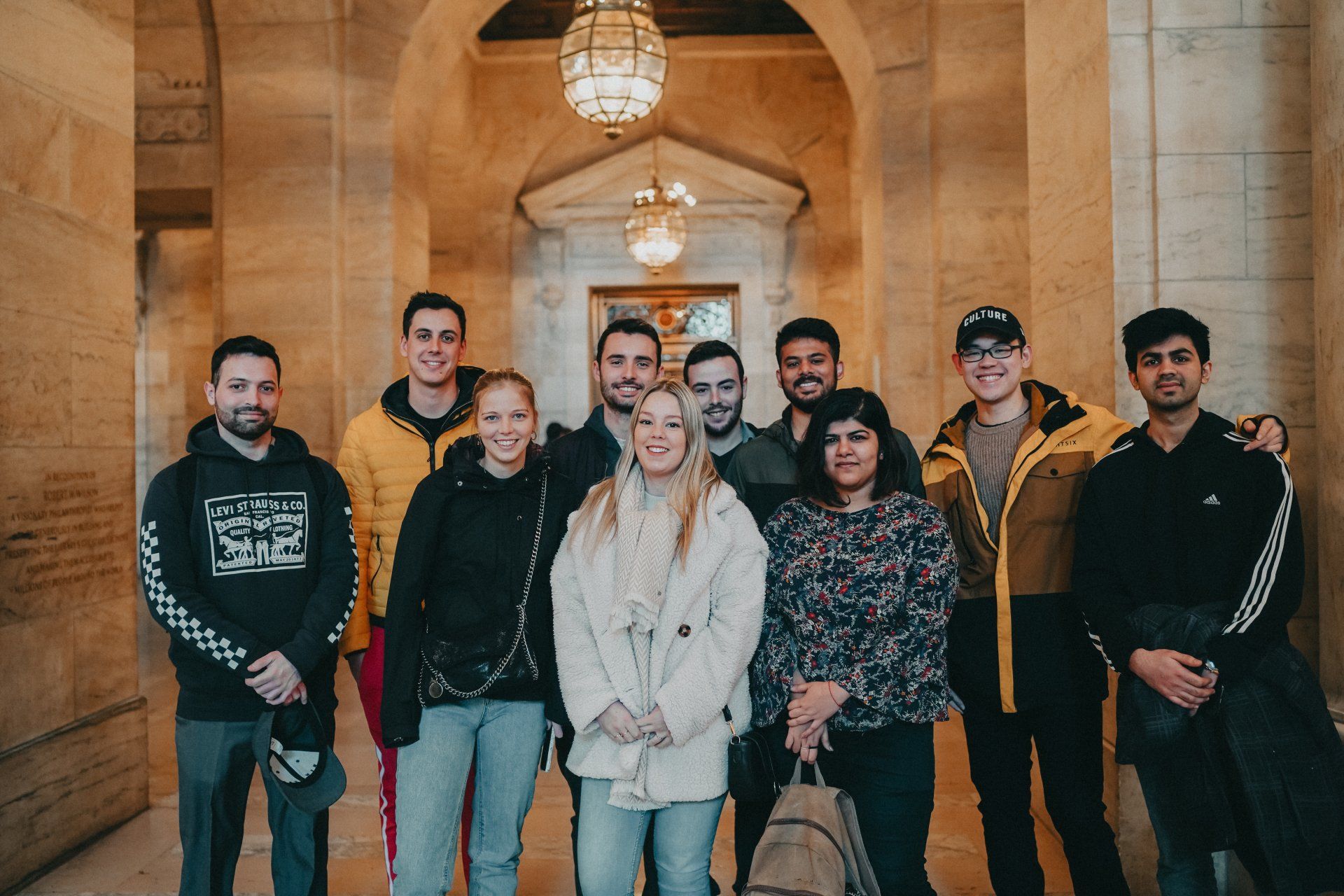 Group of nine people posing in a grand arched hallway with hanging chandeliers