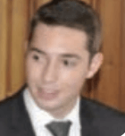 Close-up of a man in a dark suit and tie, looking slightly to the side against a wooden background