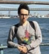 Person standing by a waterfront railing with a bridge in the background, wearing glasses and a colorful sweater.
