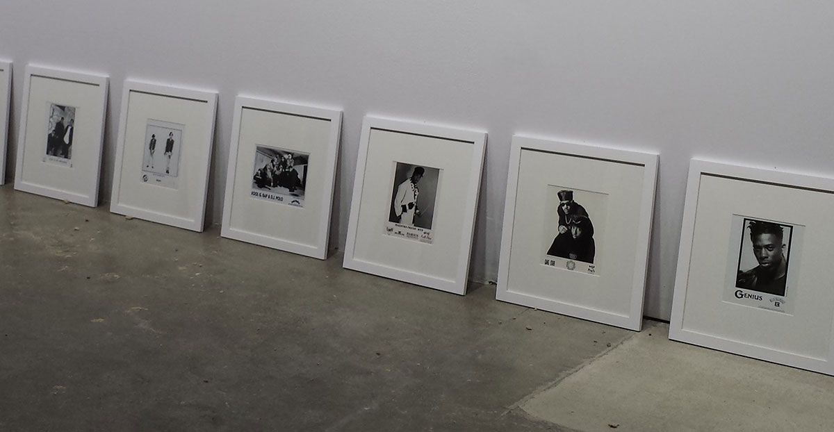 Seven framed black and white photographs leaning against a white wall on a concrete floor.