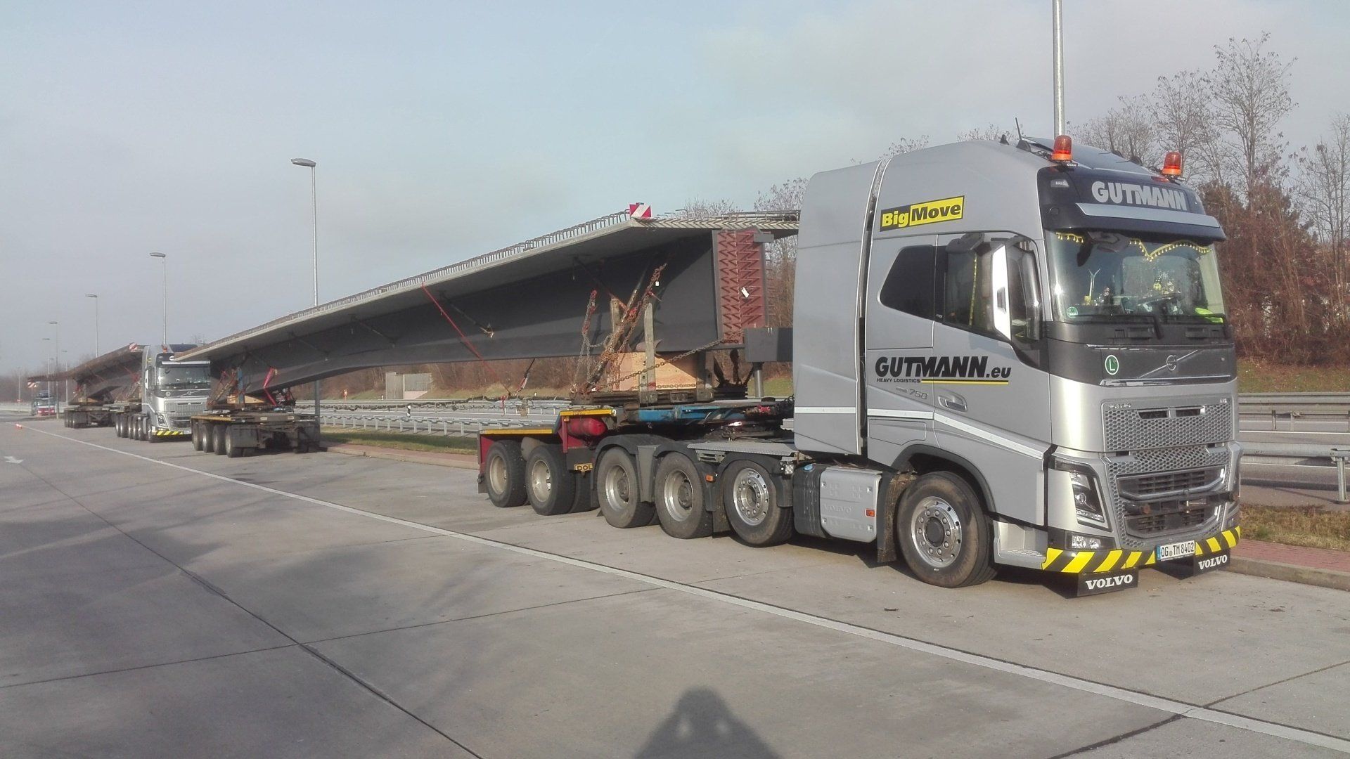 Ein silberner Sattelzug transportiert eine lange Pritschenladung auf einer Straße in einem Industriegebiet.