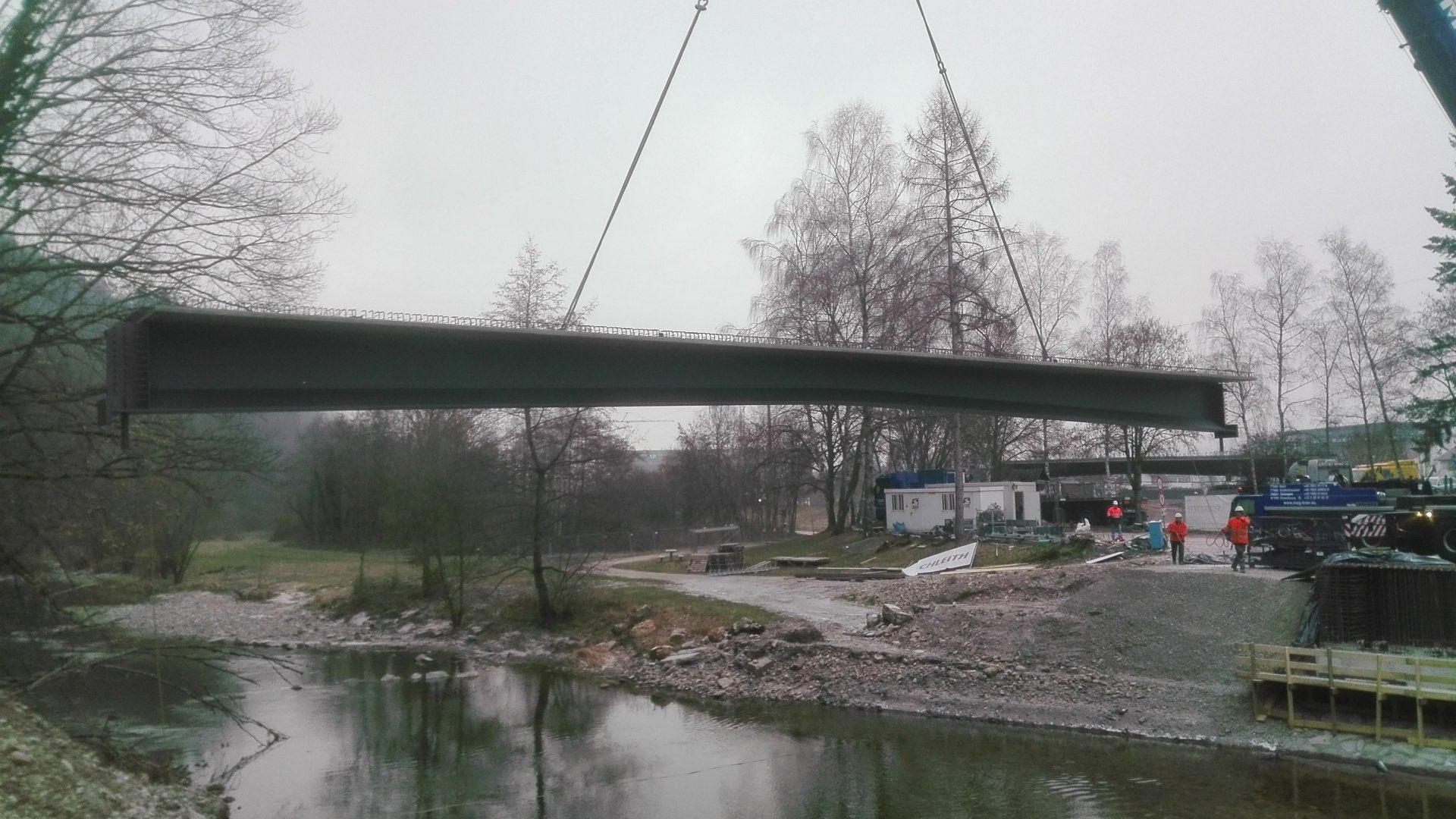 Eine dunkle Brücke überspannt einen kleinen Fluss neben einer schlammigen Baustelle und Bäumen unter einem bedeckten Himmel