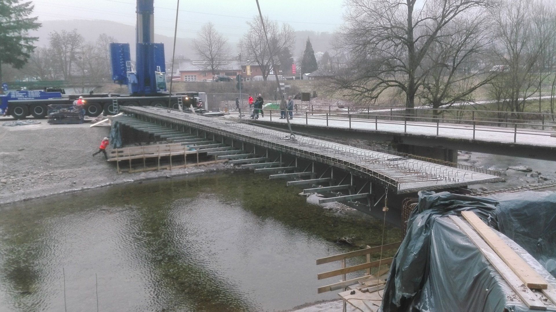 Ein verschneiter Kanal mit einer schmalen Brücke, einem blauen Kranwagen und Menschen am gegenüberliegenden Ufer