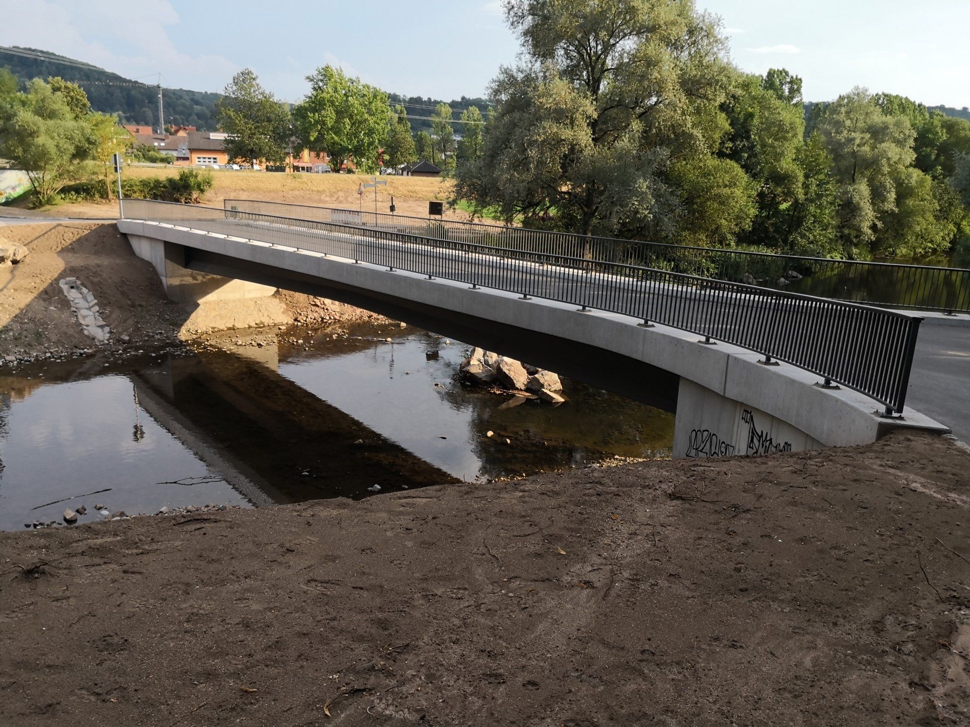 Brücke über einen ruhigen Bach mit Erdwällen, Bäumen und einer sonnigen ländlichen Landschaft.