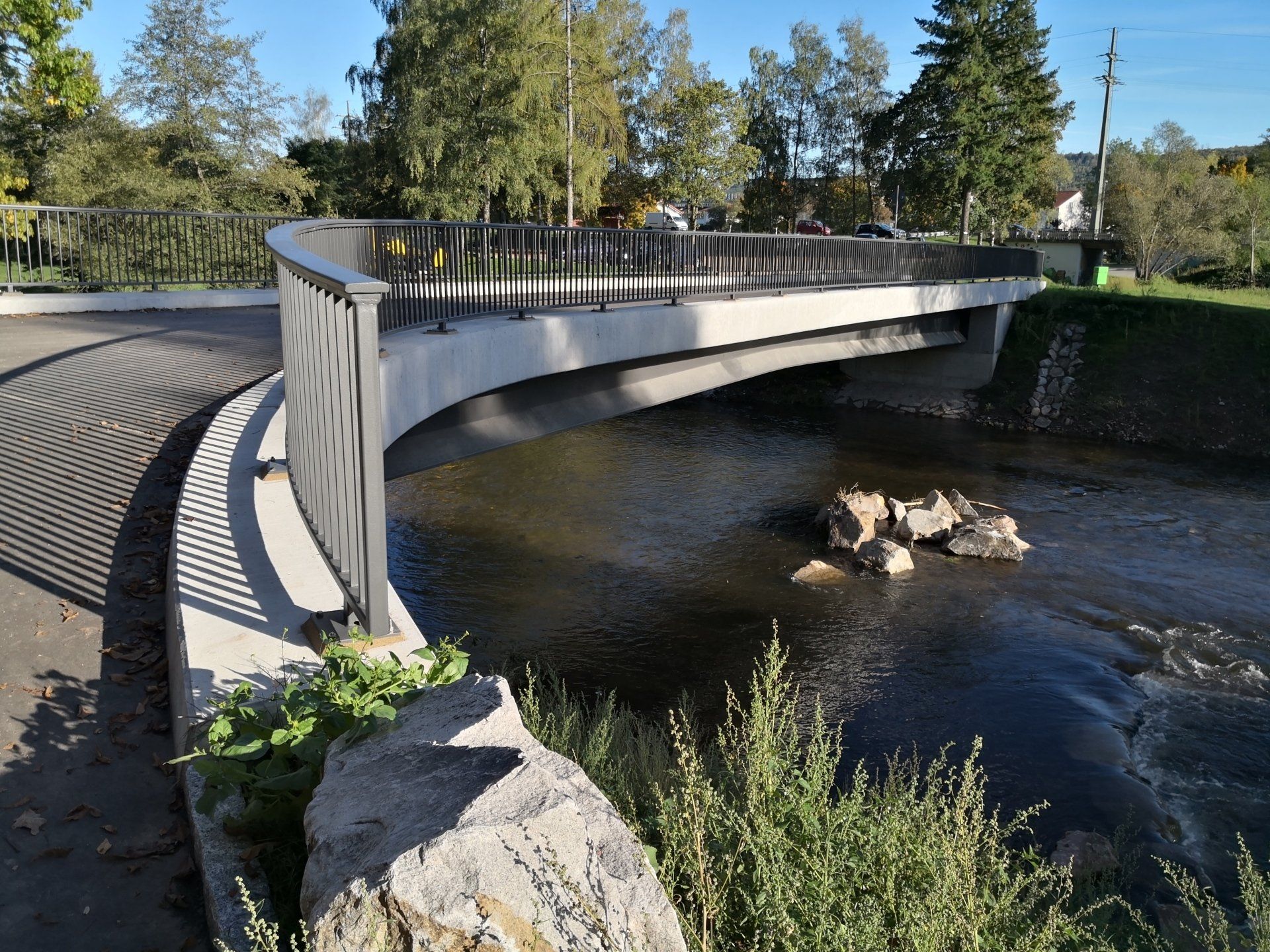 Gebogene Fußgängerbrücke über einen Bach in einem Park, mit Bäumen und Sonnenlicht, das sich im Wasser spiegelt
