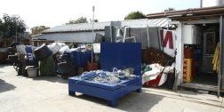 Una discarica all'aperto con rottami metallici, un tavolo blu ricoperto di detriti e un container aperto.