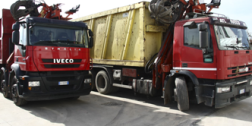 Due camion Iveco rossi con un rimorchio giallo per cassonetti parcheggiati in strada, uno dei quali carico di rottami metallici.