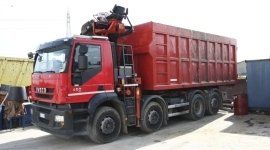 Un camion ribaltabile rosso è parcheggiato all'aperto in un cortile, carico di merci e con una gru sul tetto.