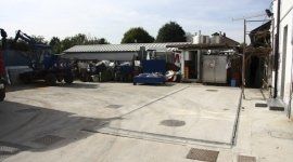 Spazio aperto in cemento con veicoli parcheggiati, un'auto blu e piccoli edifici sotto un cielo luminoso