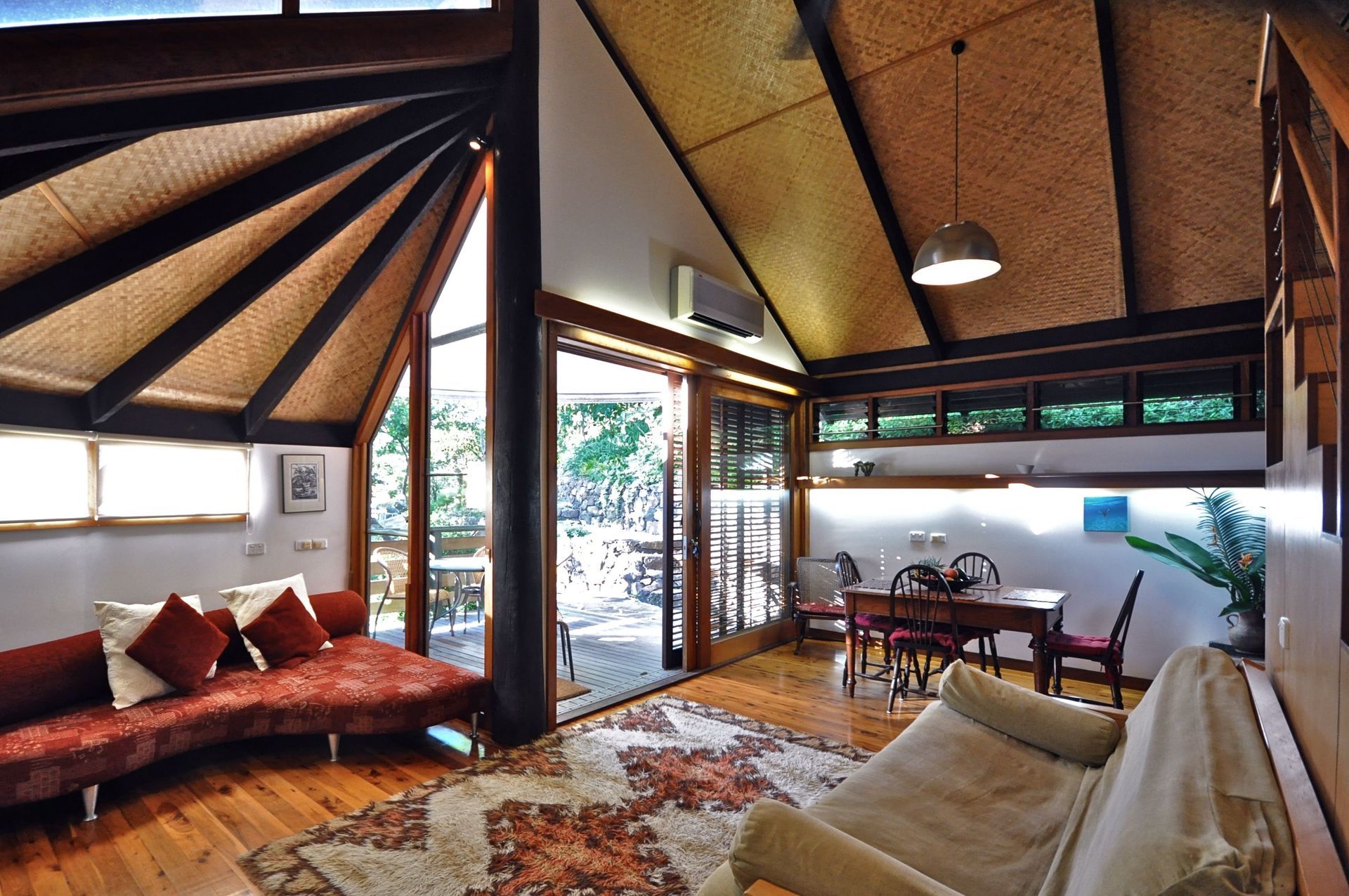 Spacious tropical living room with thatched ceiling, open glass doors, red sofa, and patterned rug.