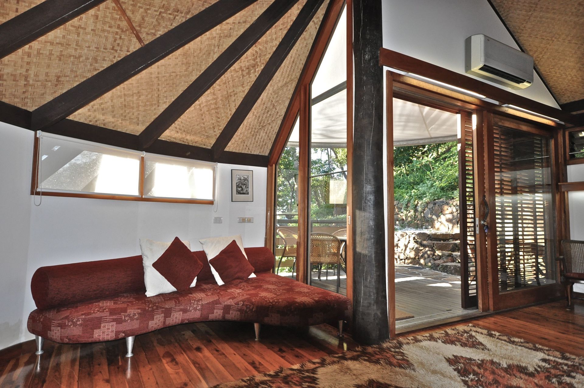 Cozy living room with a red sofa, wood-beamed ceiling, and large glass doors opening to greenery