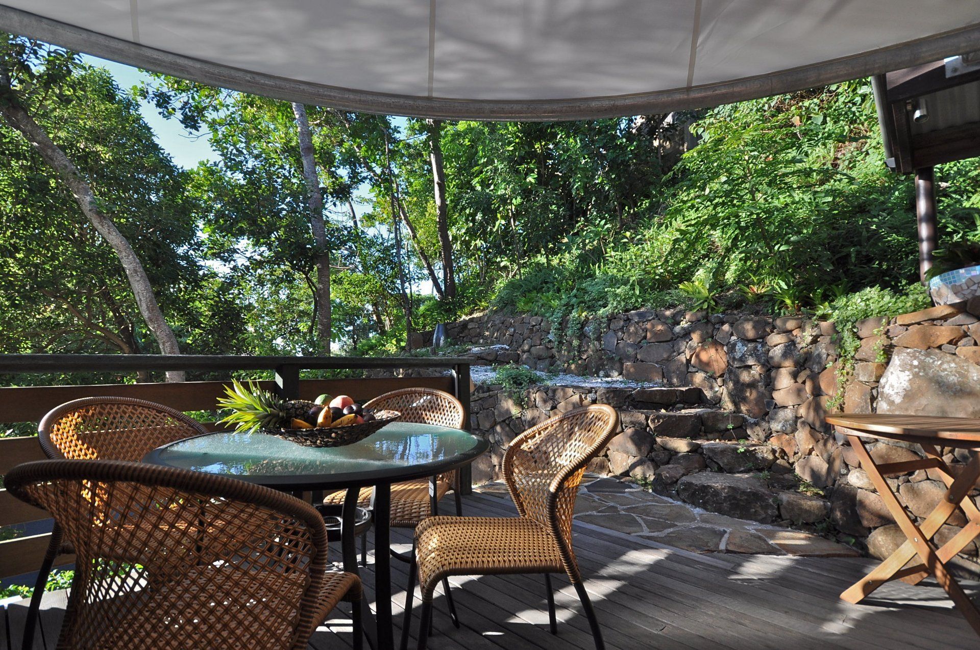 Shaded outdoor patio with wicker chairs, a glass table, and lush trees beyond a stone wall.