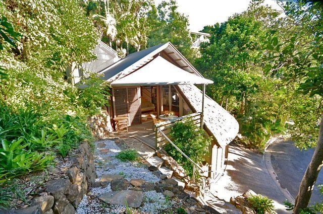 Small wooden pavilion on a stone path in a lush green garden, with a curved walkway and trees вокруг it