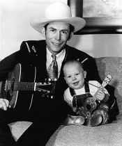 A man is sitting on a couch holding a guitar and a baby is holding a guitar.