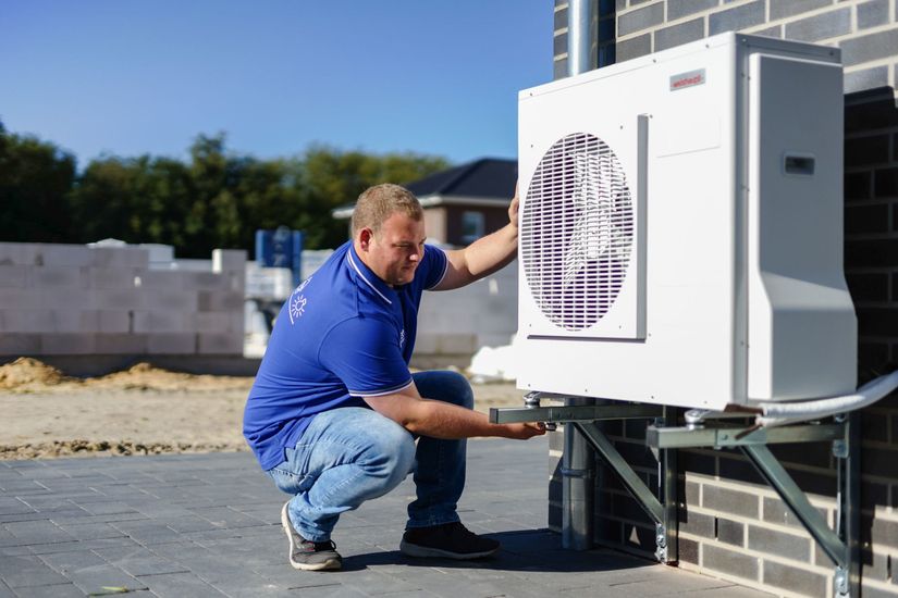 Ein Mann kniet neben einer weißen Klimaanlage auf einer Backsteinmauer.