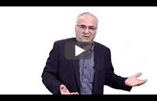 Man with glasses and blazer gesturing while speaking in front of white background.