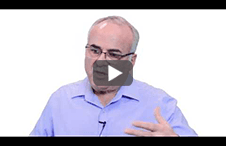 Man in blue shirt, glasses, gesturing with hands, speaking against white background.