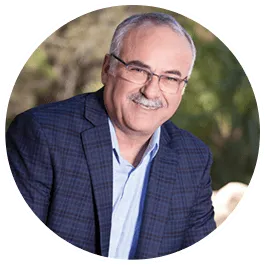 Man with glasses smiling, wearing a blue blazer and light blue shirt, outdoors.