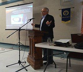 Man giving a presentation at a podium, gesturing with his hands. A projector screen and a flag are behind him.