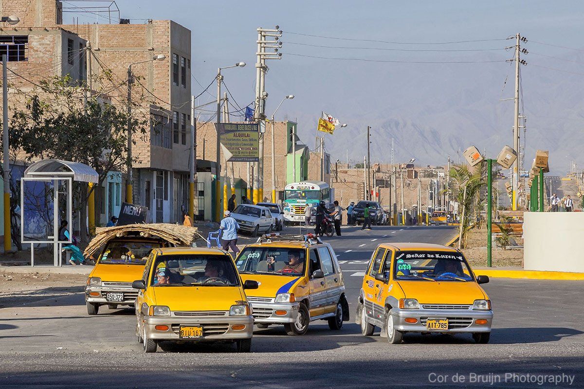 Yellow taxis on a sunlit street in a town, with buildings and mountains in the background.